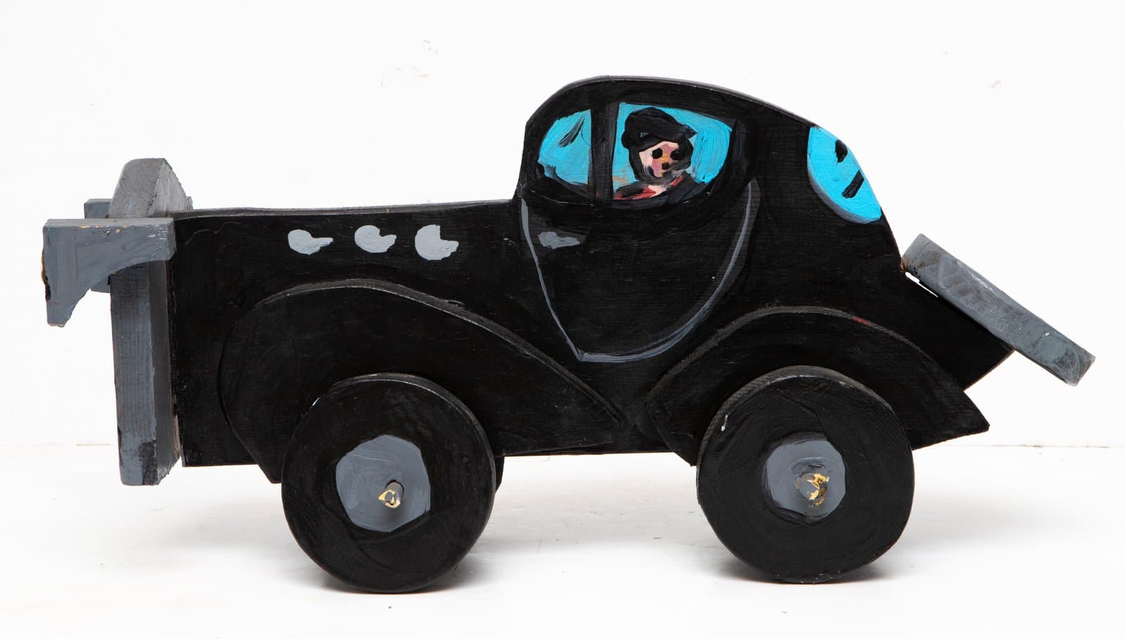 Painted wood sculpture of a black car with four wheels, blue windows, a figure inside, and gray bumpers on each end.