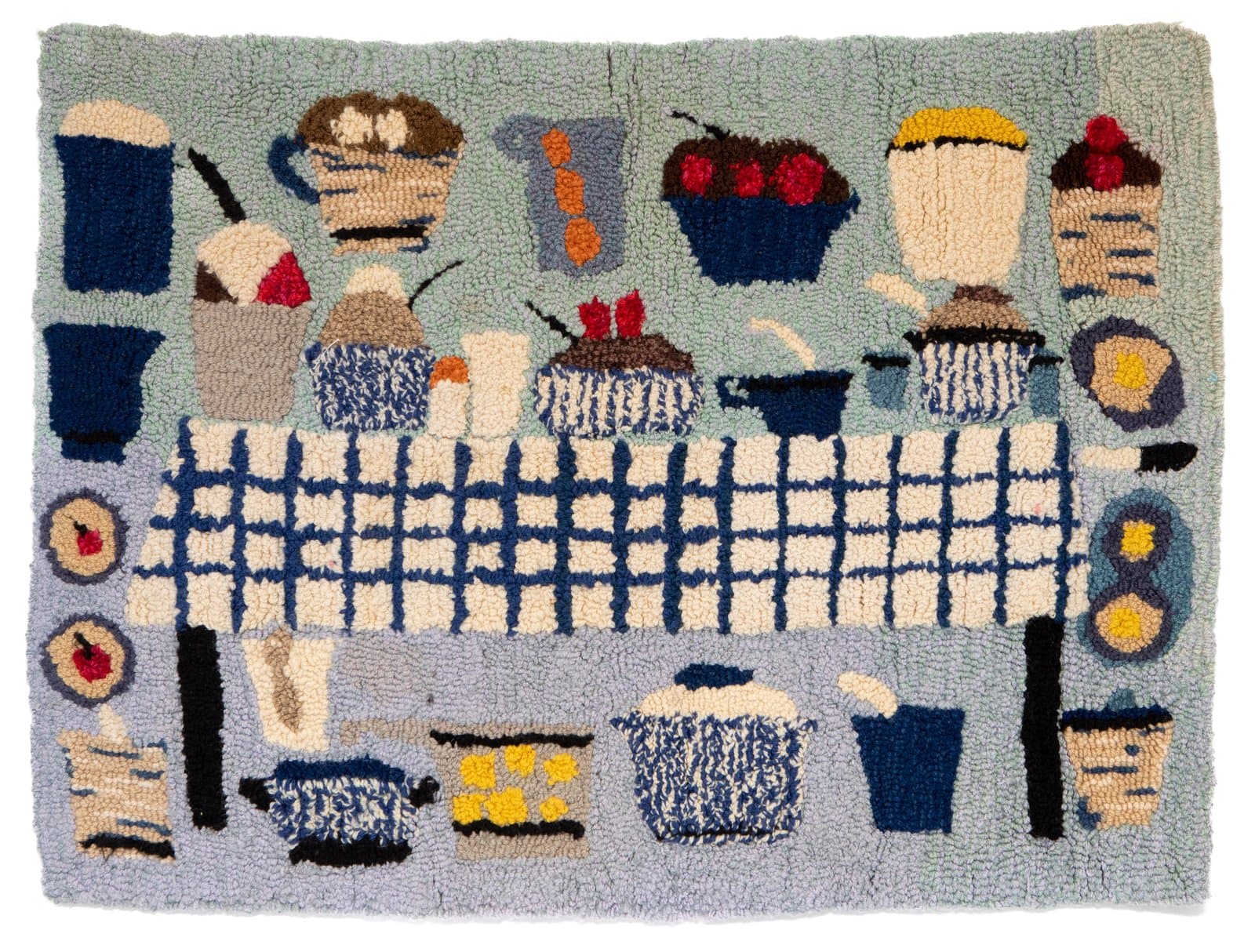 Hooked rug depicting a table spread with cupcakes, mugs, and dishes in blue, white, cream, and red on a light blue ground.