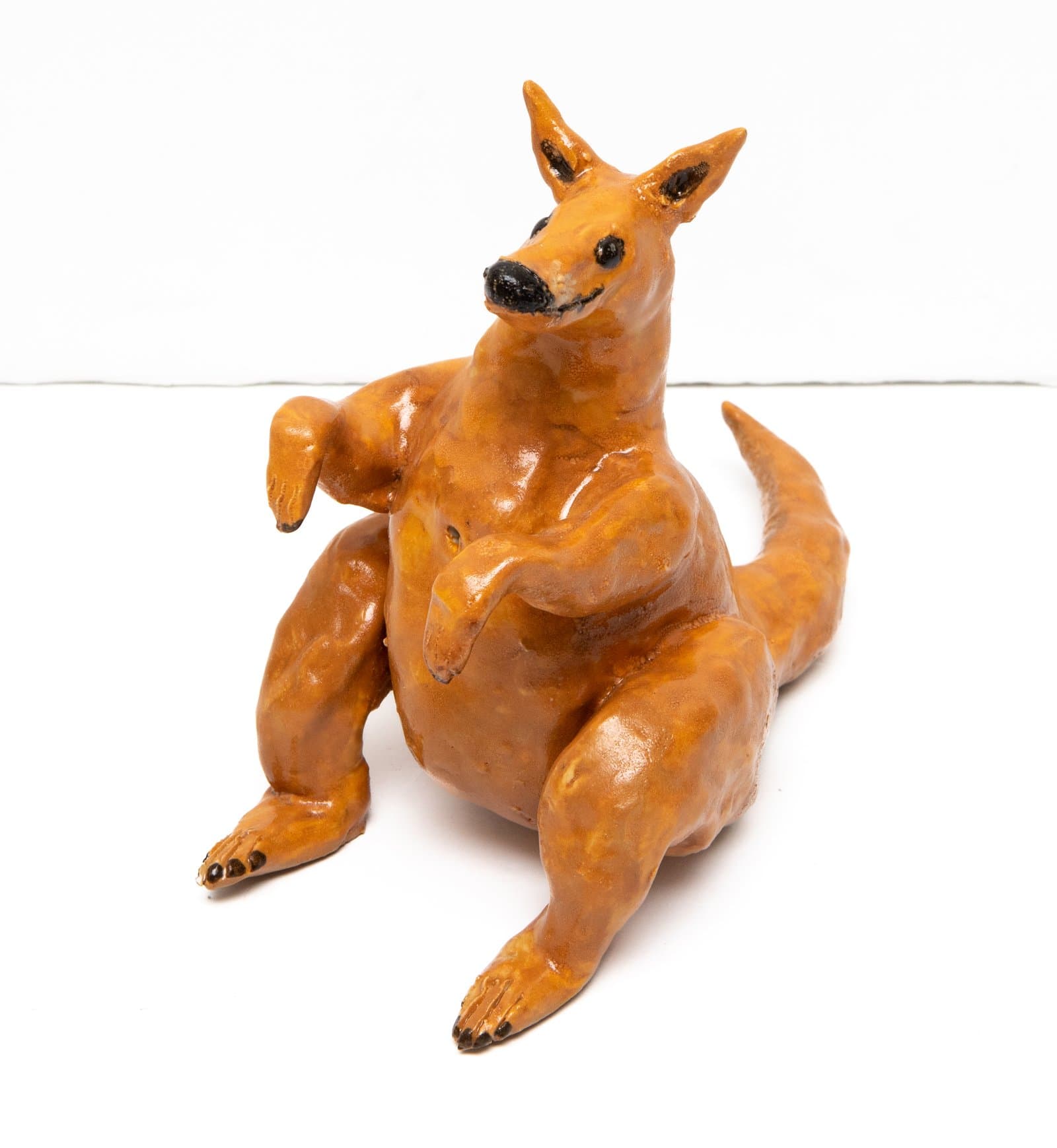 Glazed ceramic sculpture of a reclining kangaroo in warm amber-brown tones with black-accented nose, eyes, and claws.