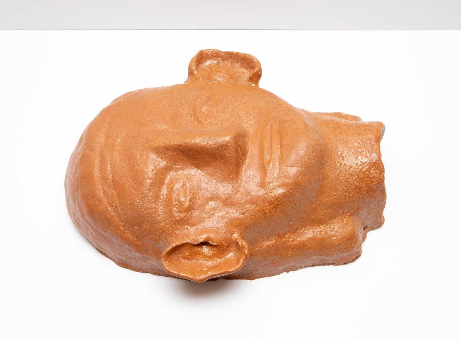 Ceramic sculpture of a reclining human head in orange-brown glaze, shown in profile against white background.