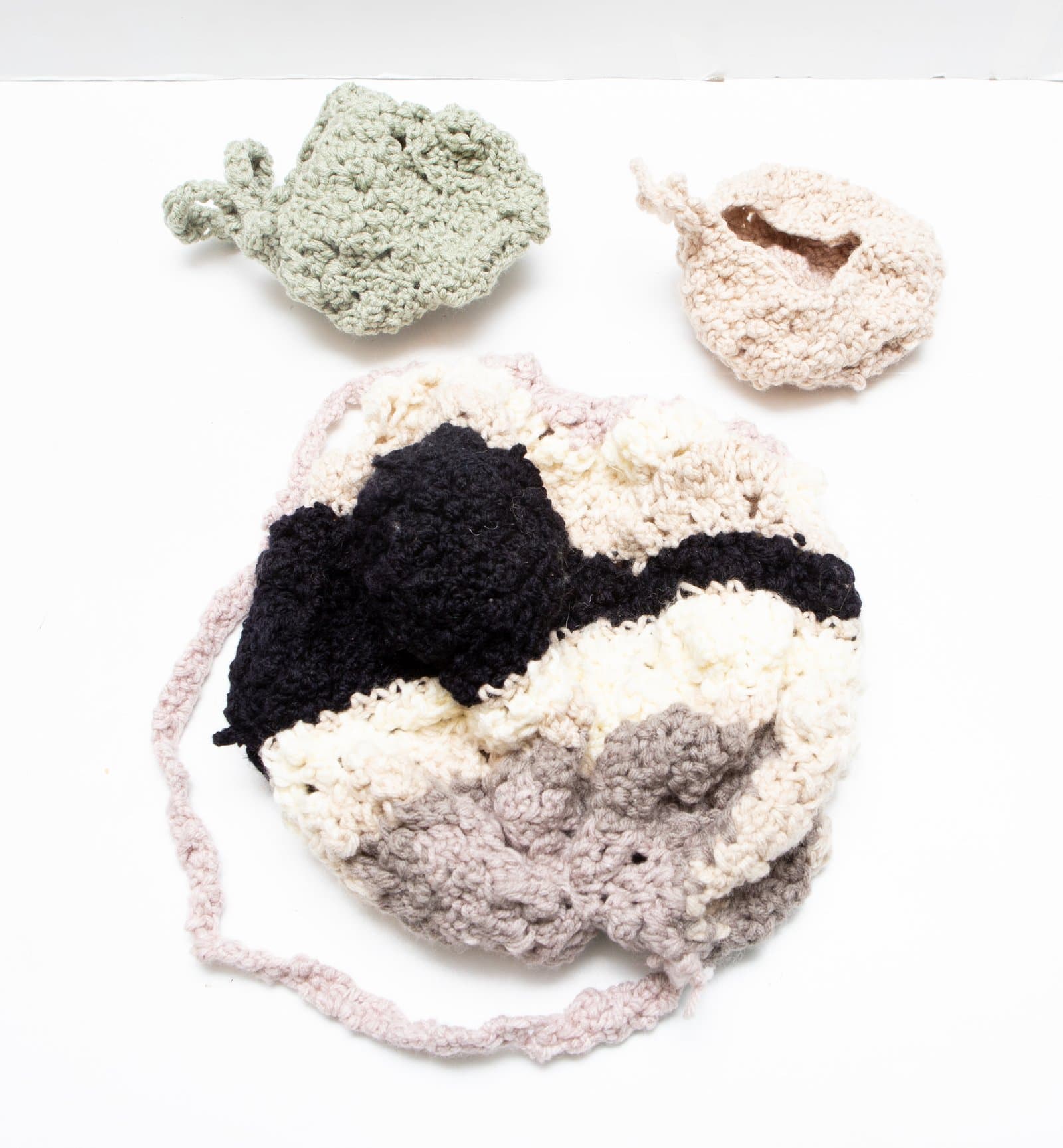 Three crocheted textile objects on white background: a large ruffled pouch, a small sage green form, and a small cream circular pouch.