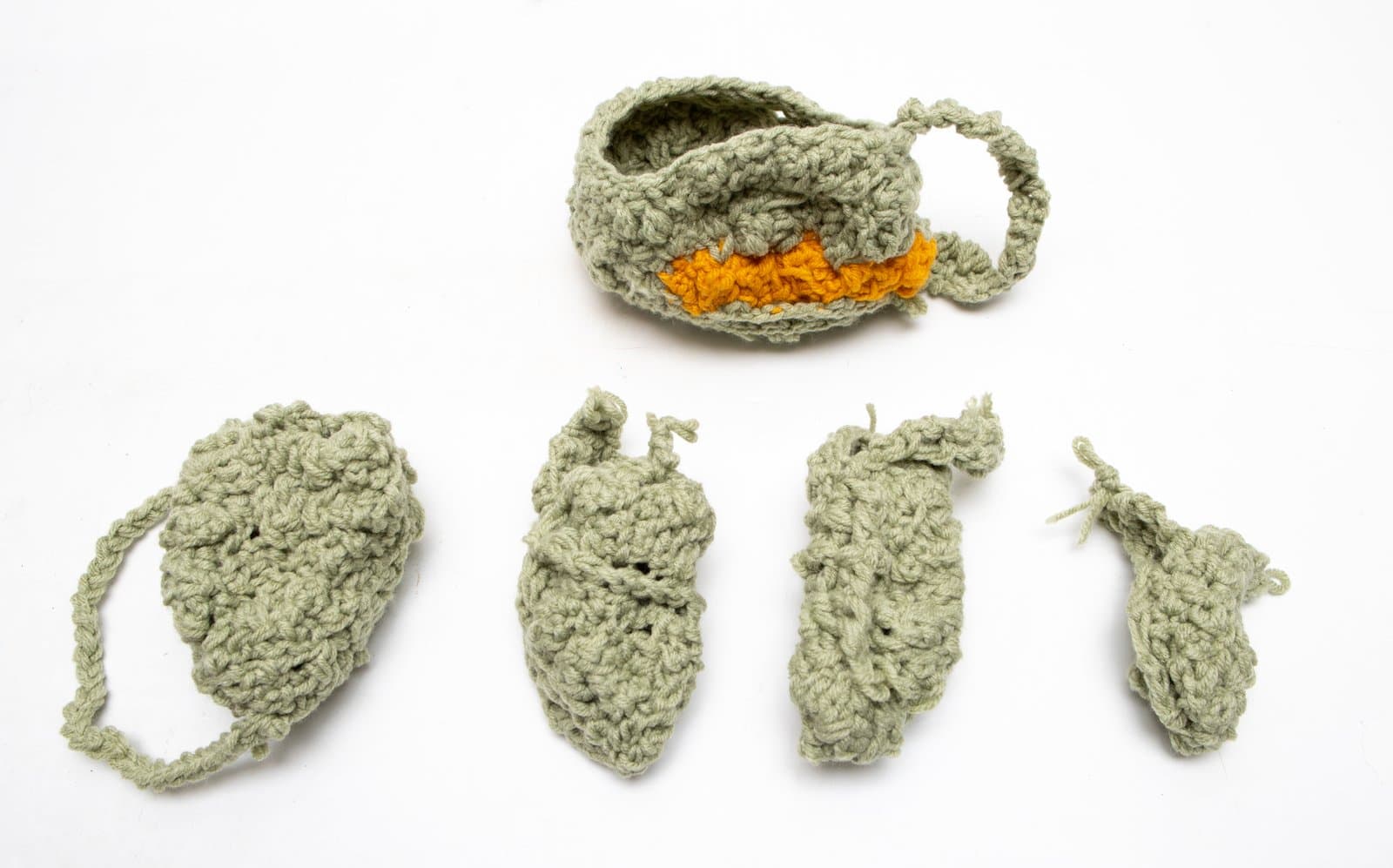 Five crocheted forms in sage green yarn arranged on white background; a cup-like vessel with orange accent and four flat leaf-shaped pieces.