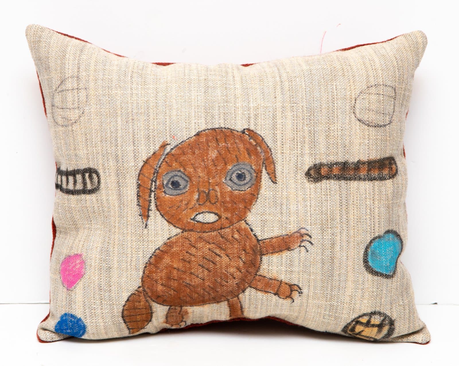 Decorative fabric pillow with hand-drawn brown creature and scattered colorful objects on beige woven textile.