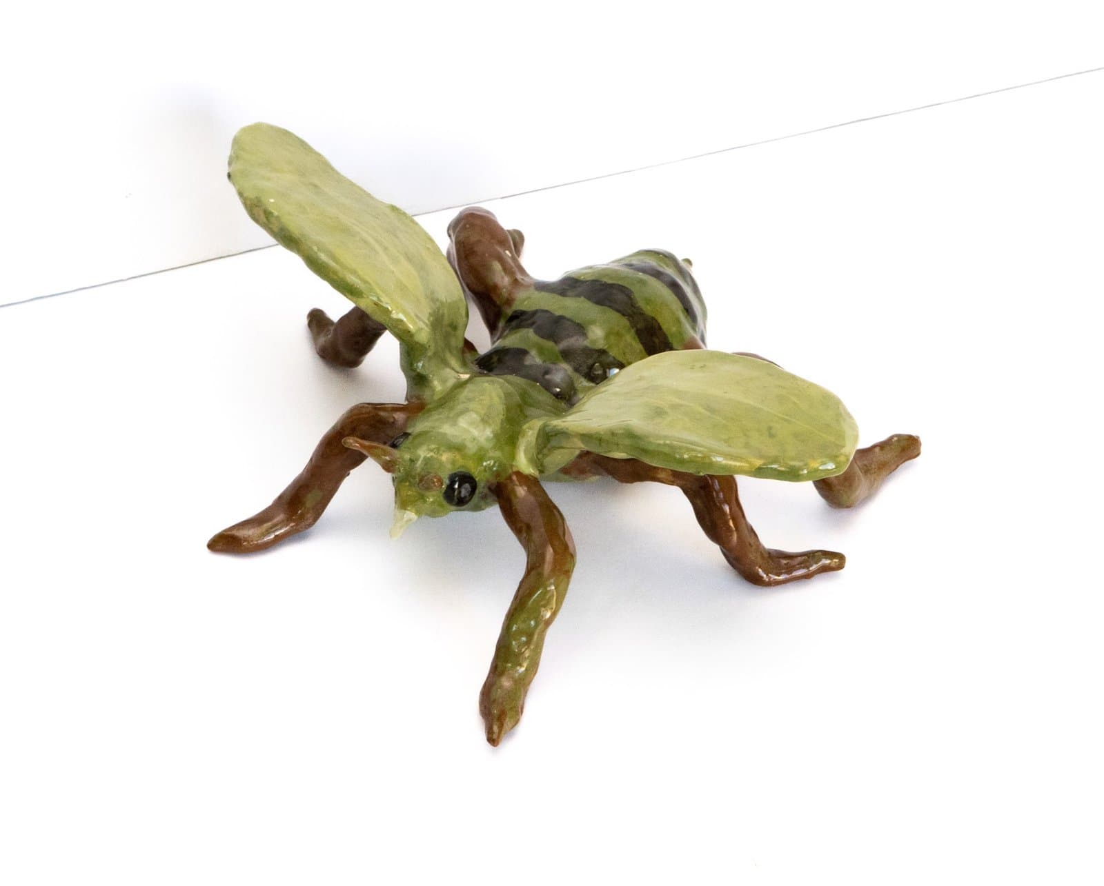 Ceramic sculpture of a green and brown grasshopper with elongated wings and six legs on white background.