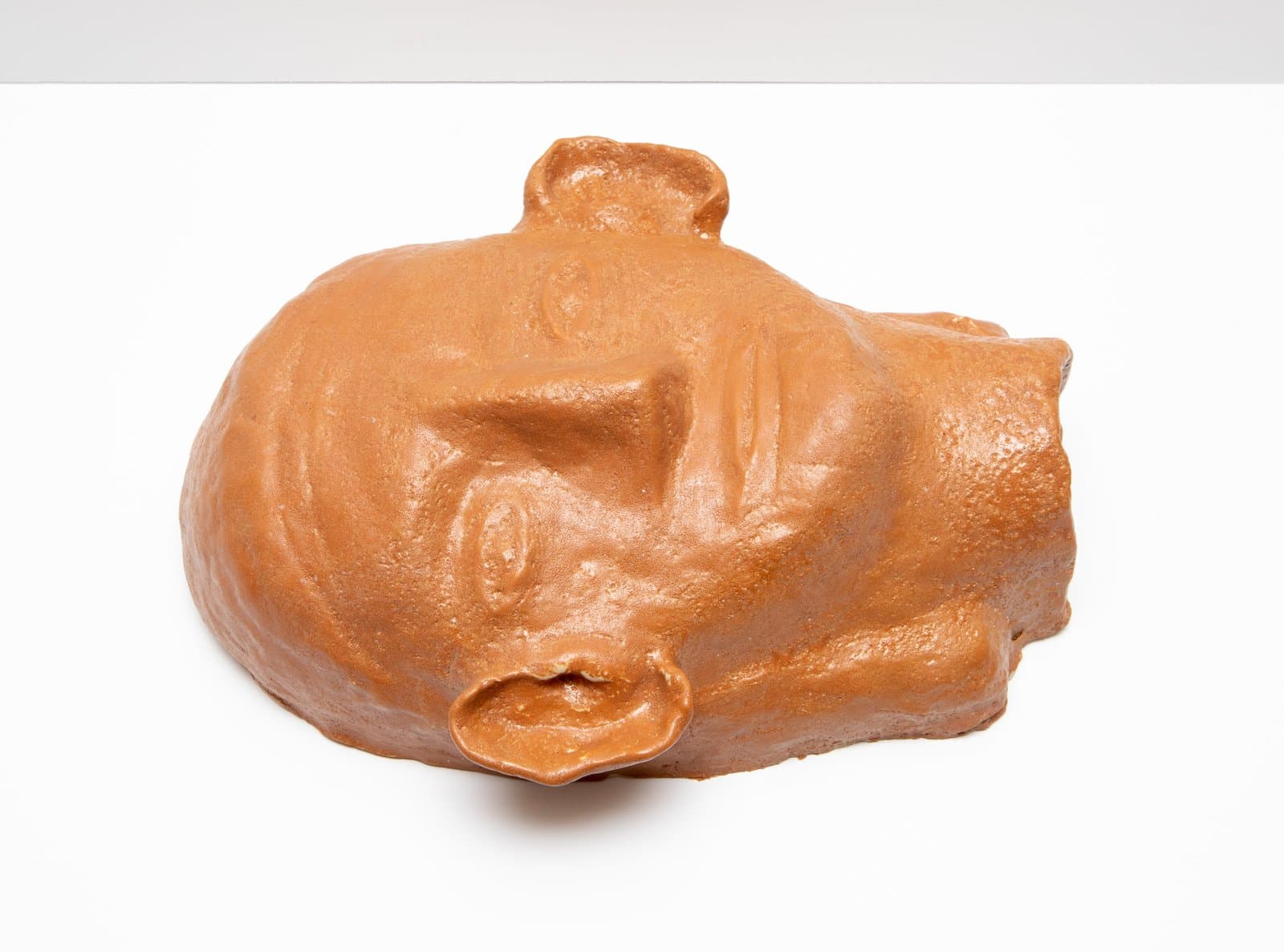 Ceramic sculpture of a human head lying on its side, rendered in terracotta-orange glaze with simplified facial features.