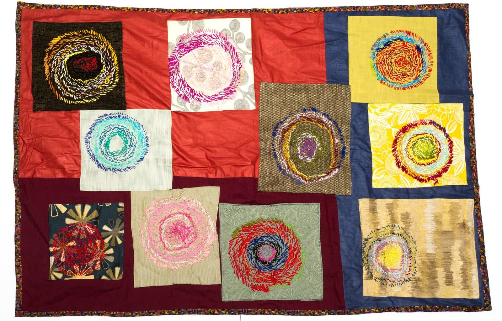 Patchwork textile artwork with ten squares featuring embroidered spiral designs in various colors on different fabric backgrounds