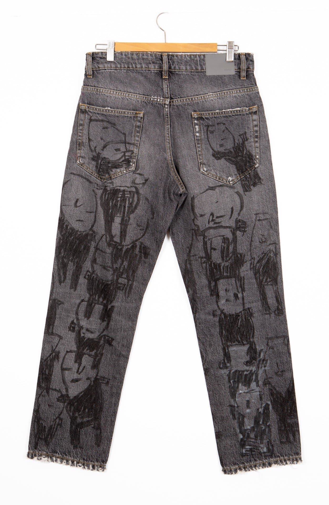Gray denim jeans with black marker drawings of figures and faces covering the entire surface, hanging on wooden hanger.