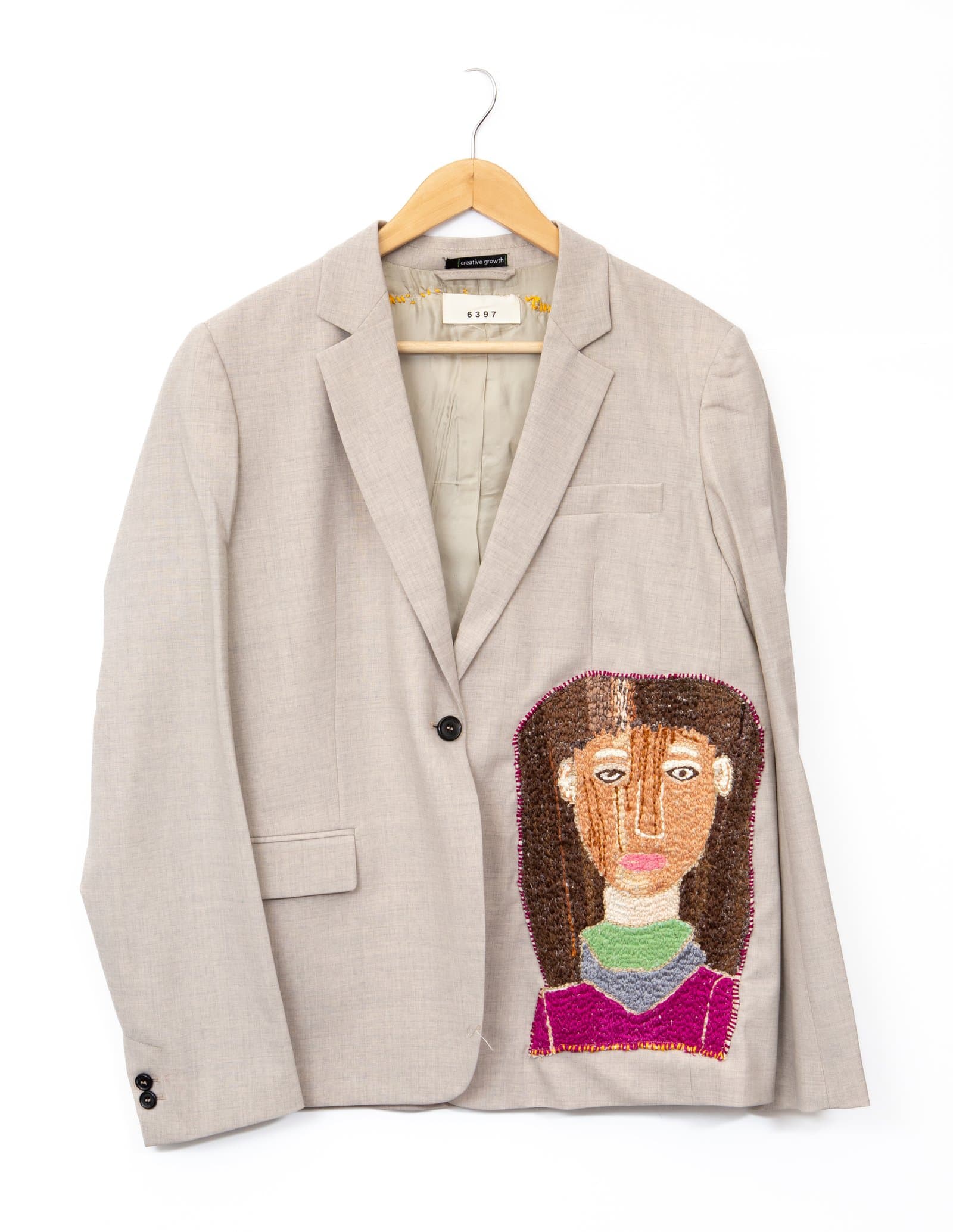 Beige blazer with a large embroidered portrait of a figure with brown hair on the right front panel, displayed on a wooden hanger.