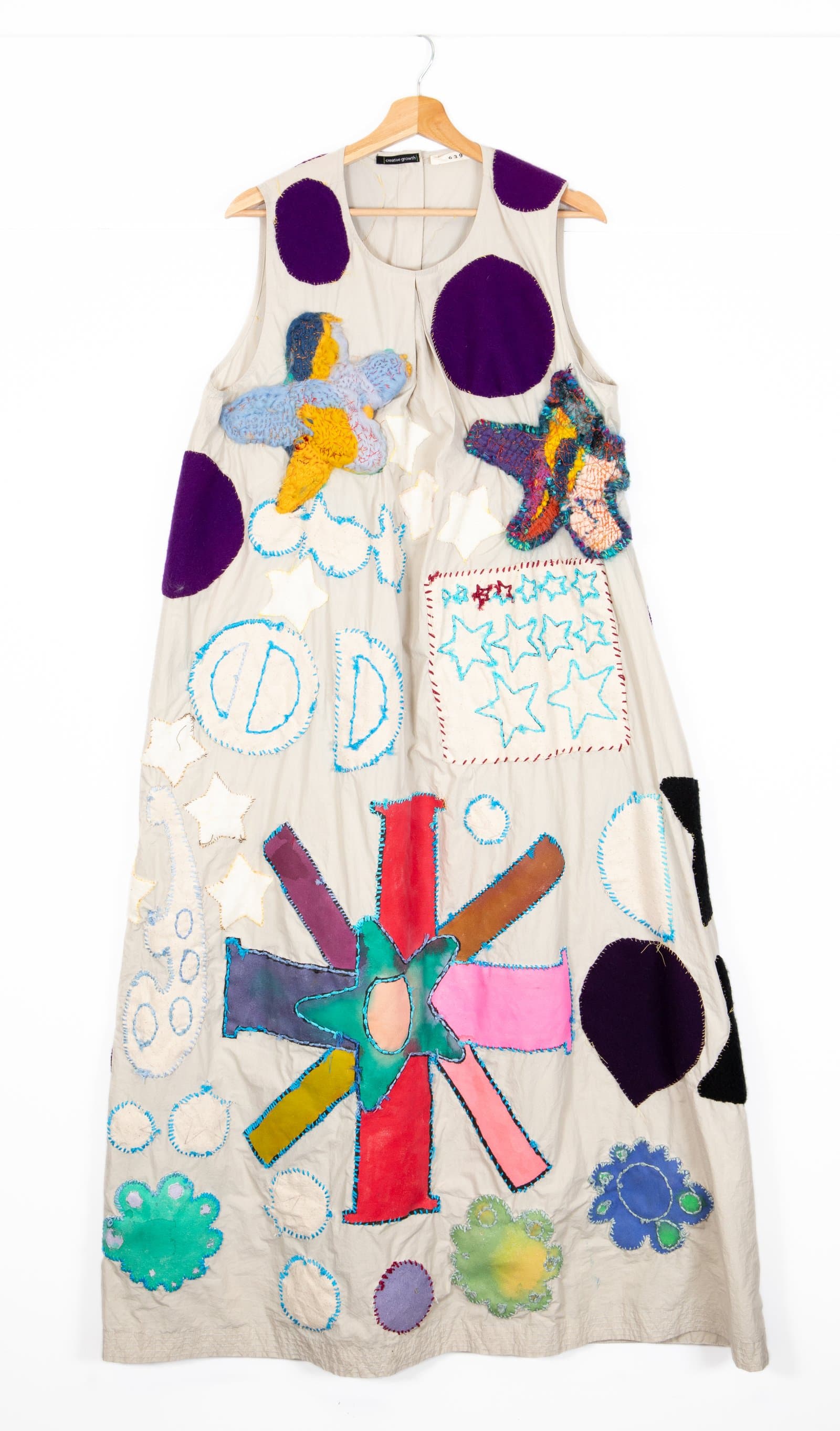 Sleeveless linen dress covered in colorful fabric appliqués and embroidery including stars, circles, and starburst shapes.