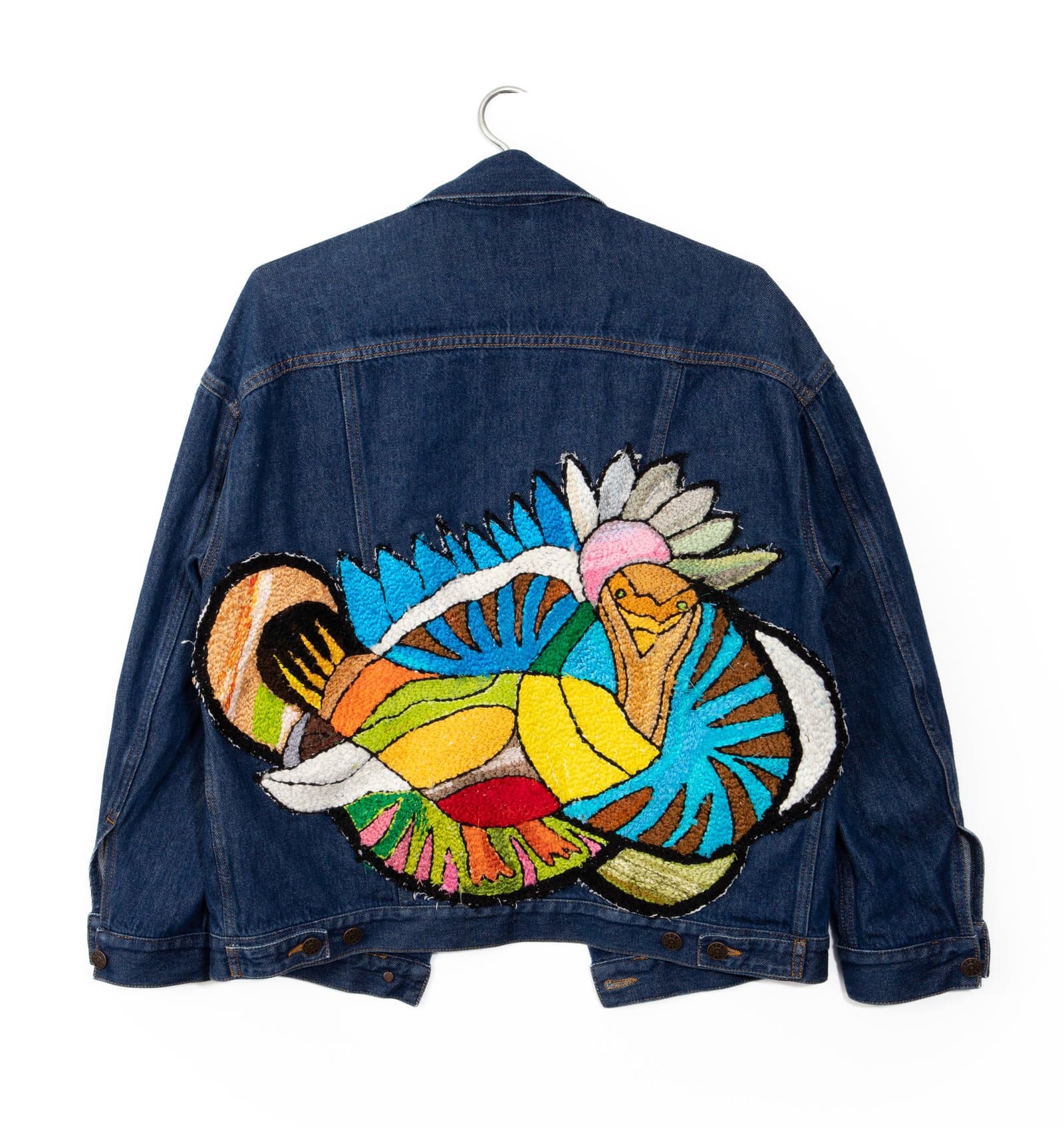 Dark denim jacket displayed on hanger, back view, with a large multicolor embroidered or tufted patch depicting an abstract bird figure.