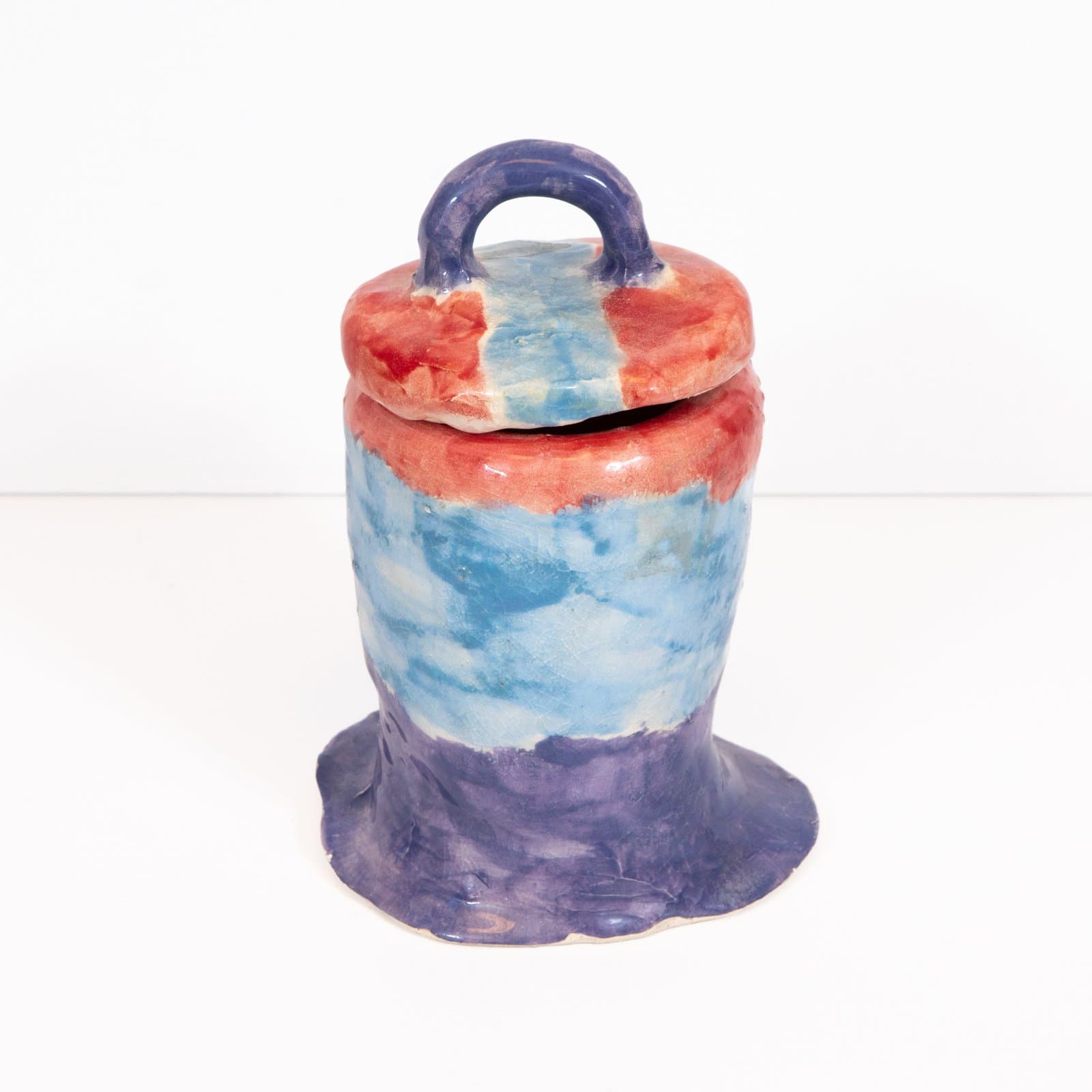 Glazed ceramic lidded jar with loop handle, featuring blended red, blue, and purple glazes and a flared, ruffled base.