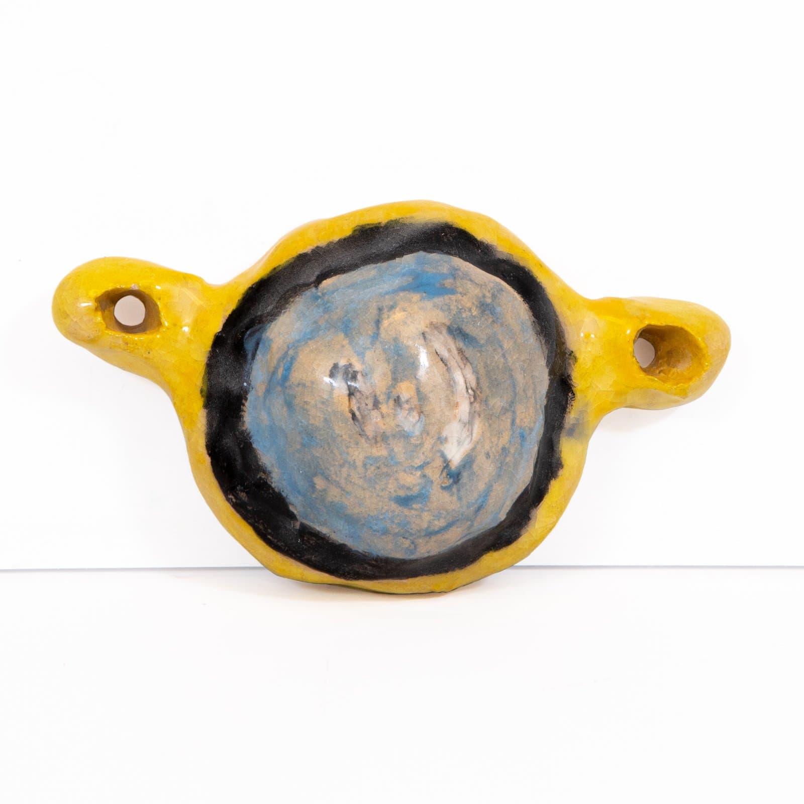 Small glazed ceramic piece with yellow outer form, black ring, and blue-tan swirled center, with two small side handles.