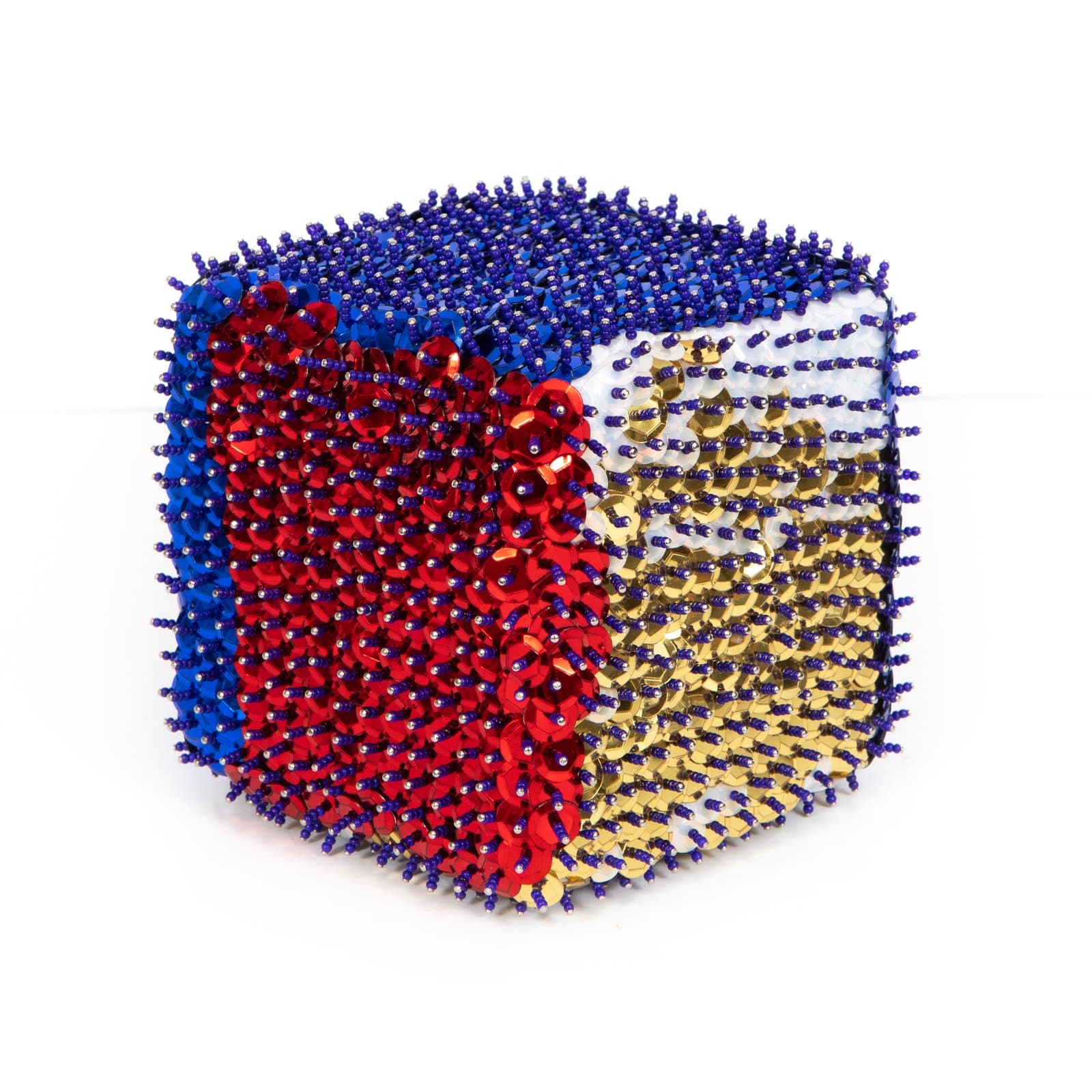 Cube sculpture densely covered in sequins and beads in red, blue, gold, and white arranged in diagonal color blocks.