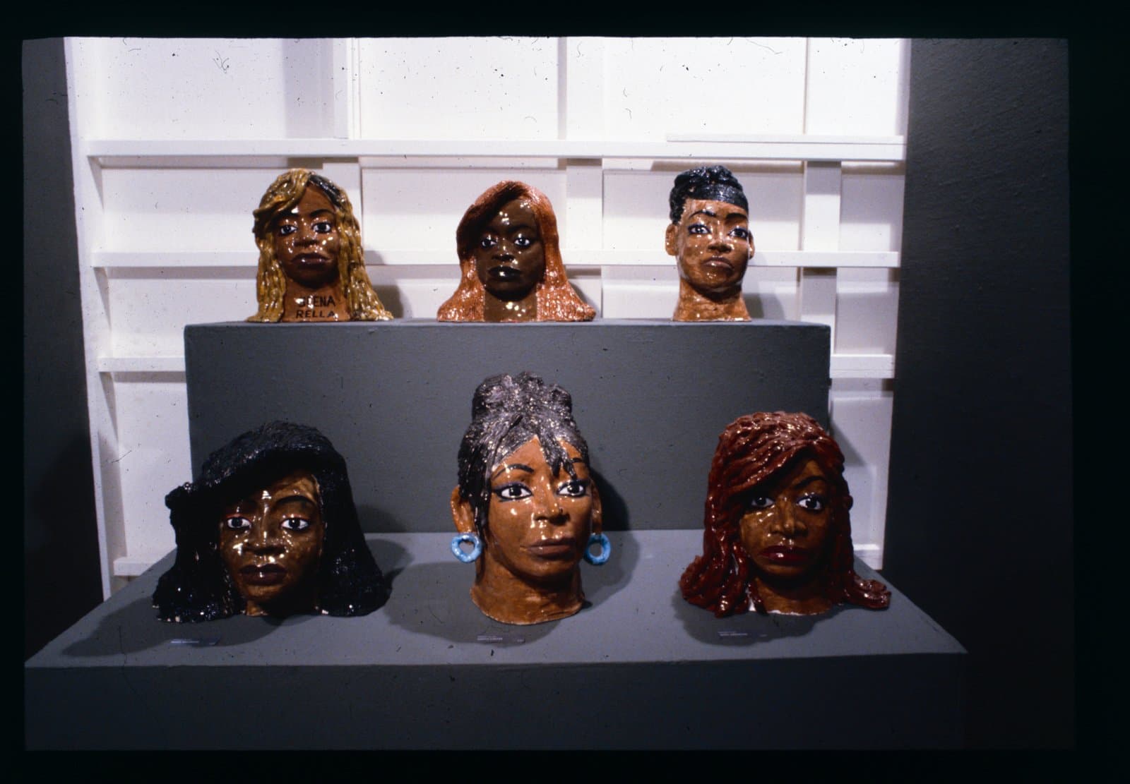Six glazed ceramic portrait busts of women with brown skin tones displayed on a two-tiered gray pedestal.