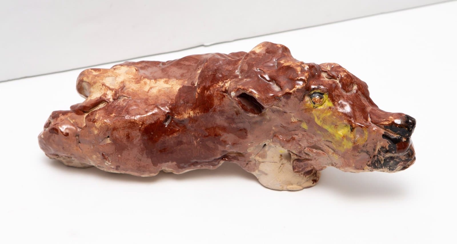 Ceramic sculpture of a reclining lion with glazed brown surface and yellow accent details on white background.