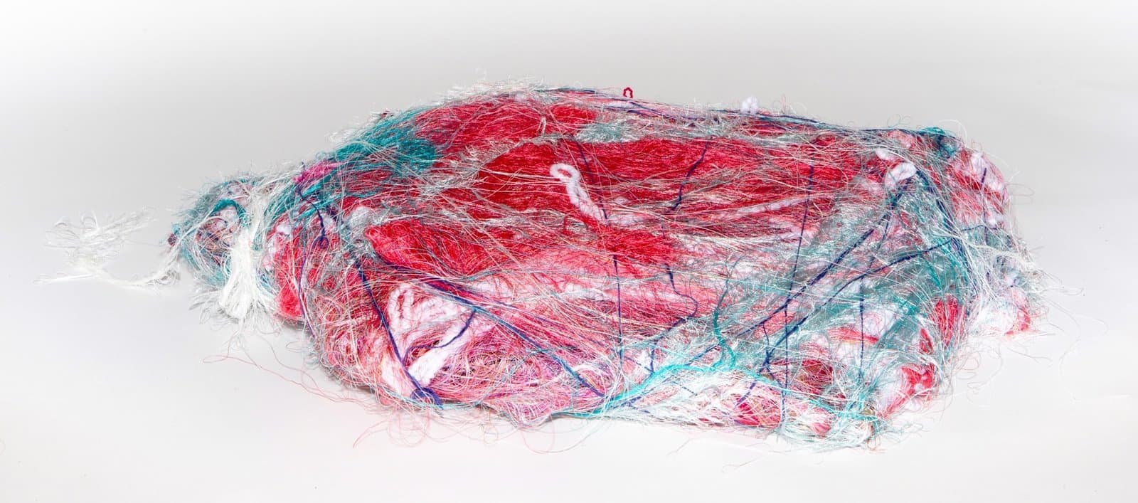Wrapped sculpture with multicolored threads covering an oval form in red, turquoise, and white on gray background.