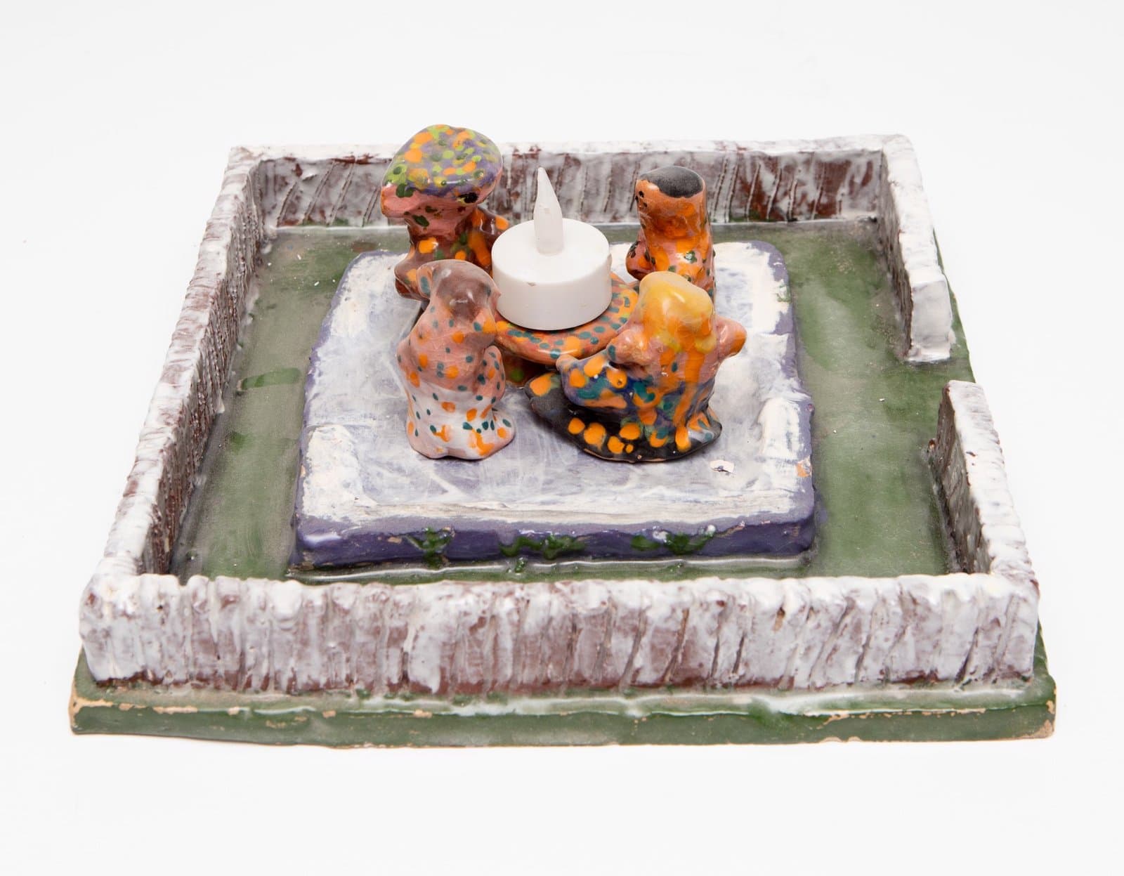 Ceramic sculpture featuring small figurines arranged around a white candle on a platform within a square basin.