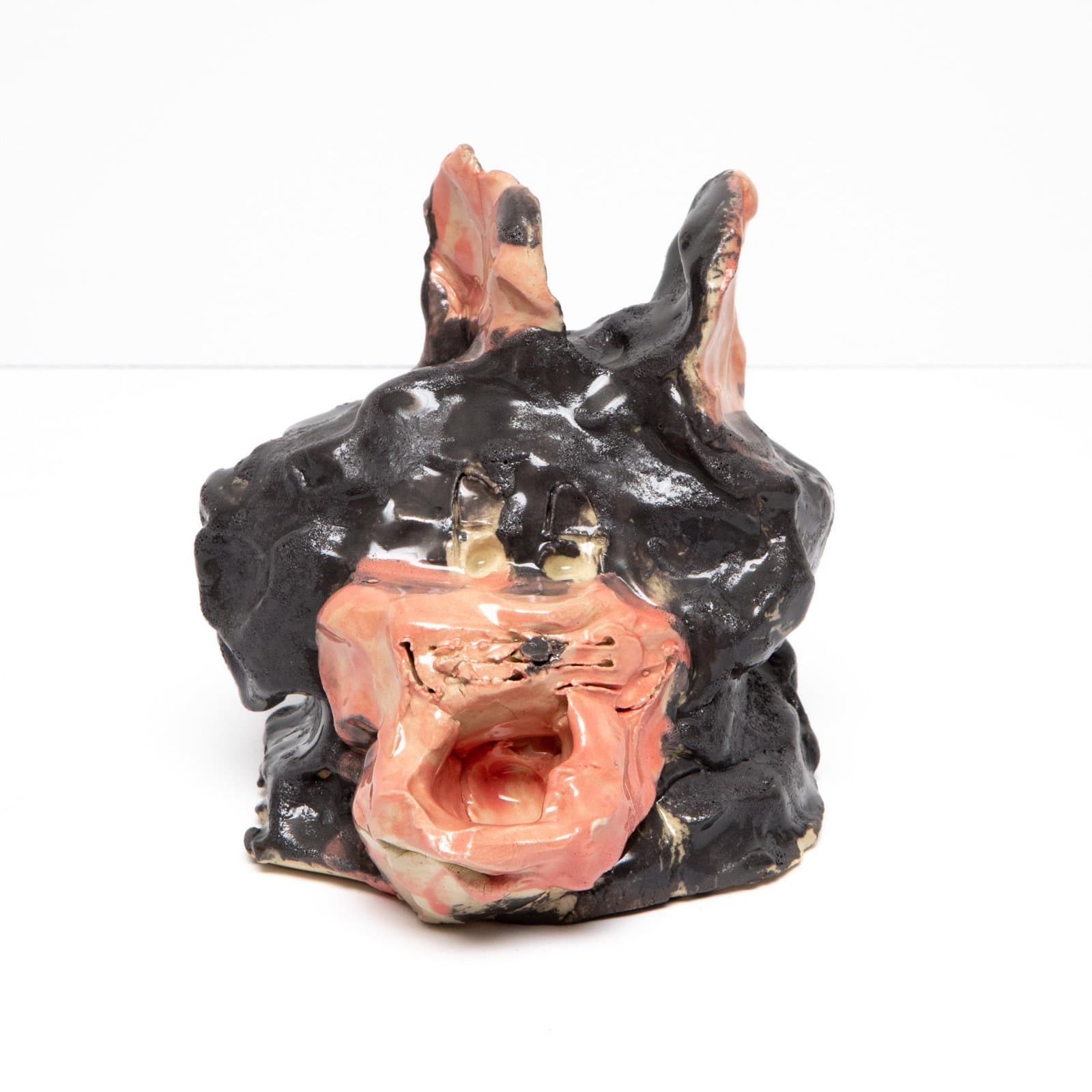 Ceramic sculpture of a grotesque face with open mouth, glazed in black and pink with protruding ears and teeth.