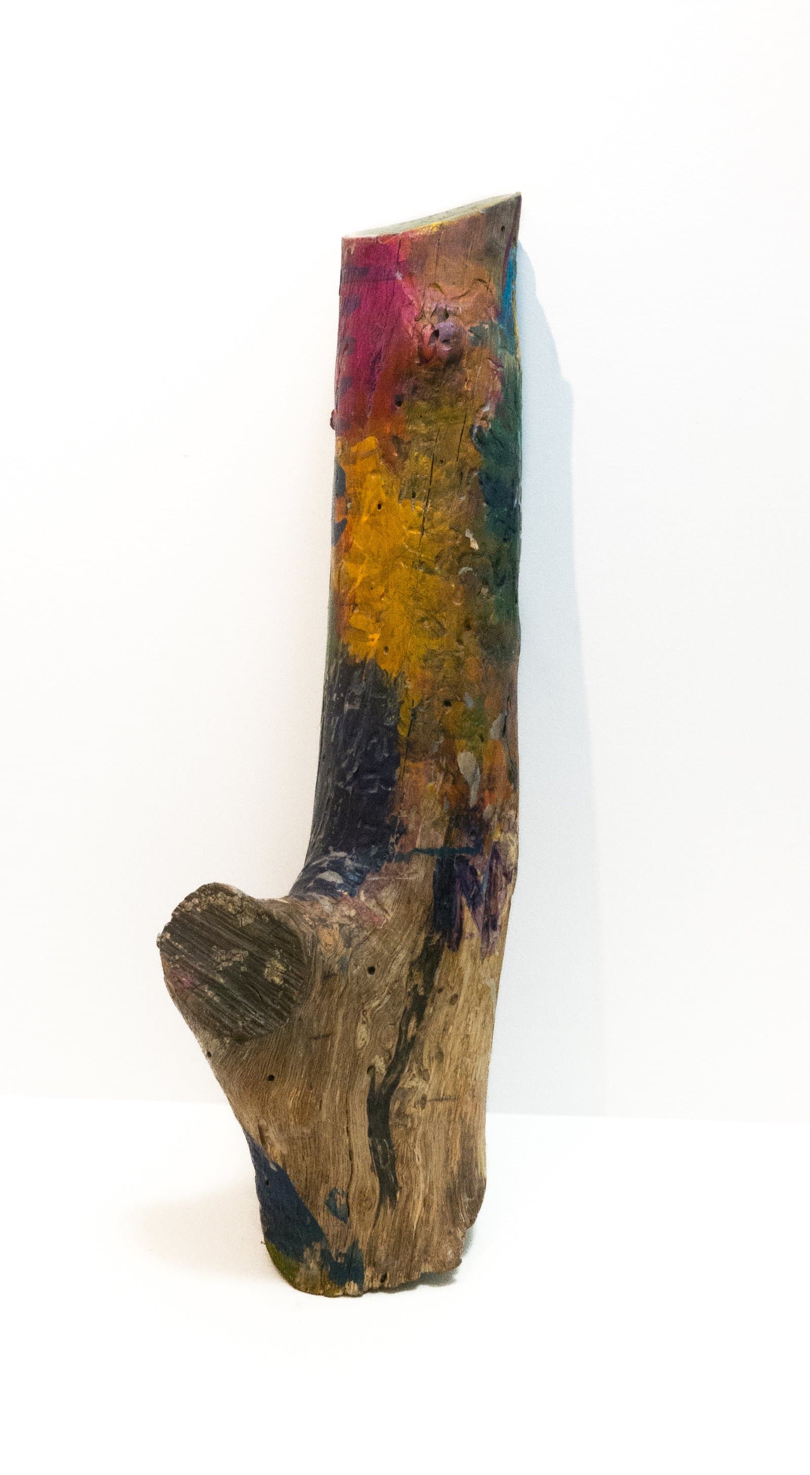 Sculptural painted wood piece; a curved tree branch or log with vibrant patches of pink, yellow, teal, and dark paint on natural wood.