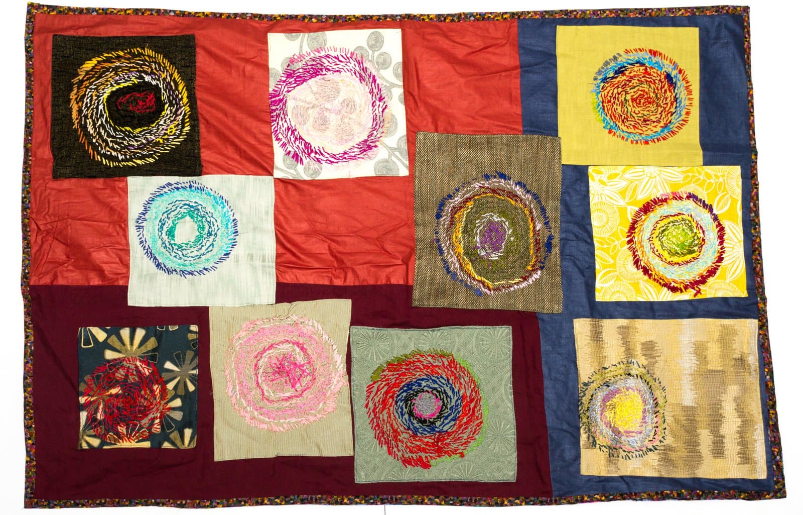 Textile quilt with ten fabric panels, each featuring a spiral or circular embroidered design in multicolored thread.