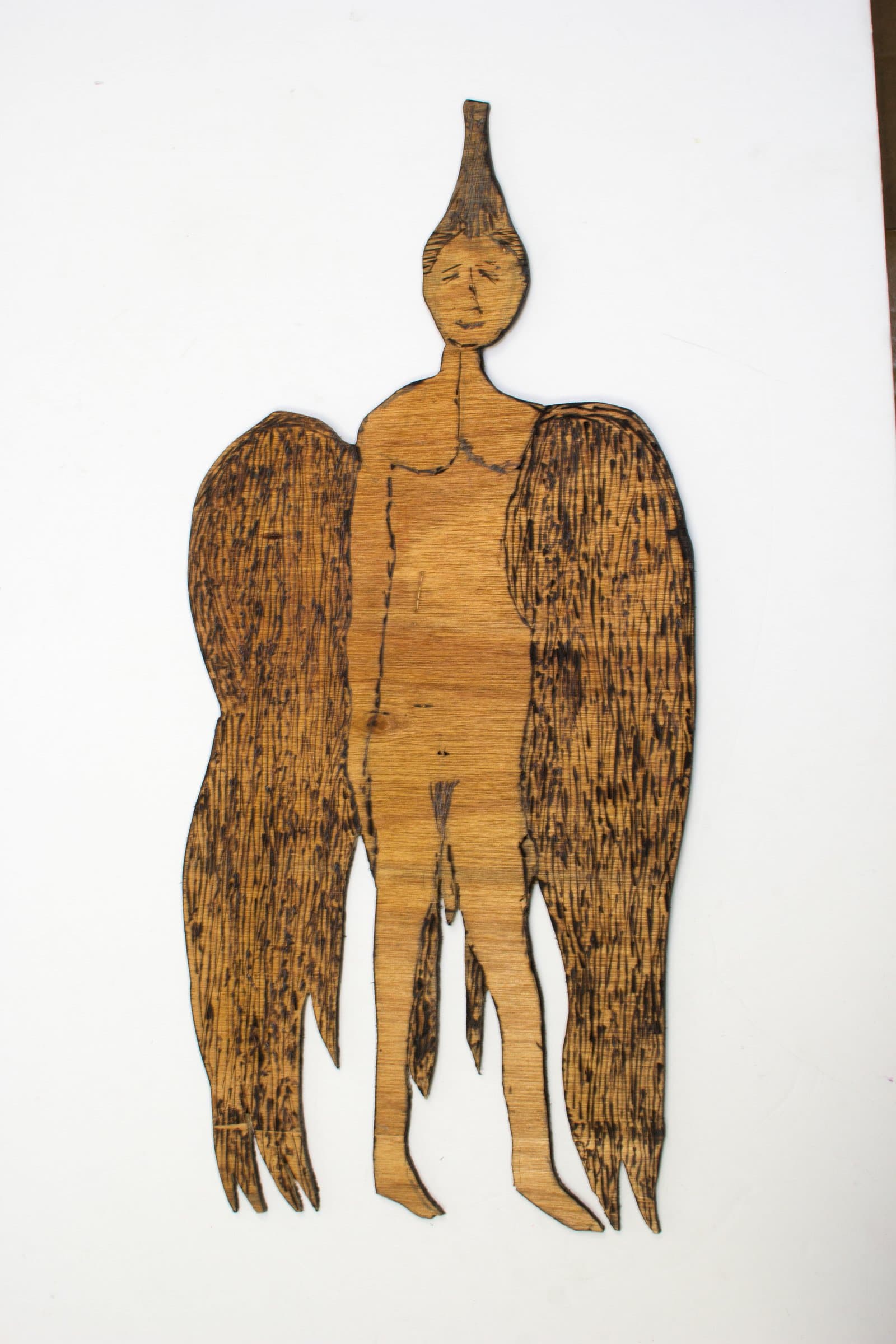 Cutout wood figure of a winged human with a pointed head, decorated with pyrography burns, mounted on white background.