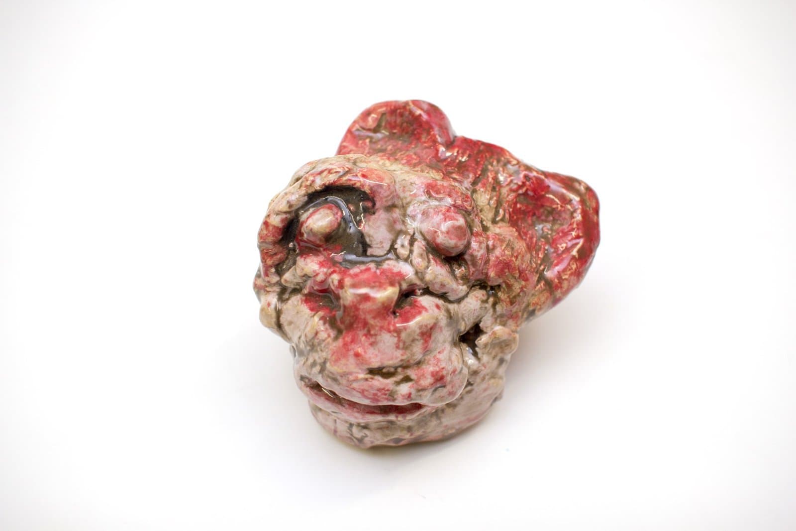 Ceramic sculpture of distorted face with red and pink glazed surface, bulbous features and hollow eye sockets