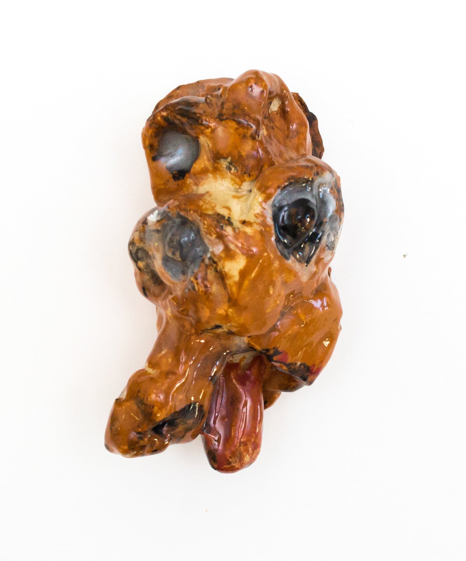 Small glazed ceramic sculpture of a dog face with bulging eyes and protruding tongue, in amber, brown, and dark gray.