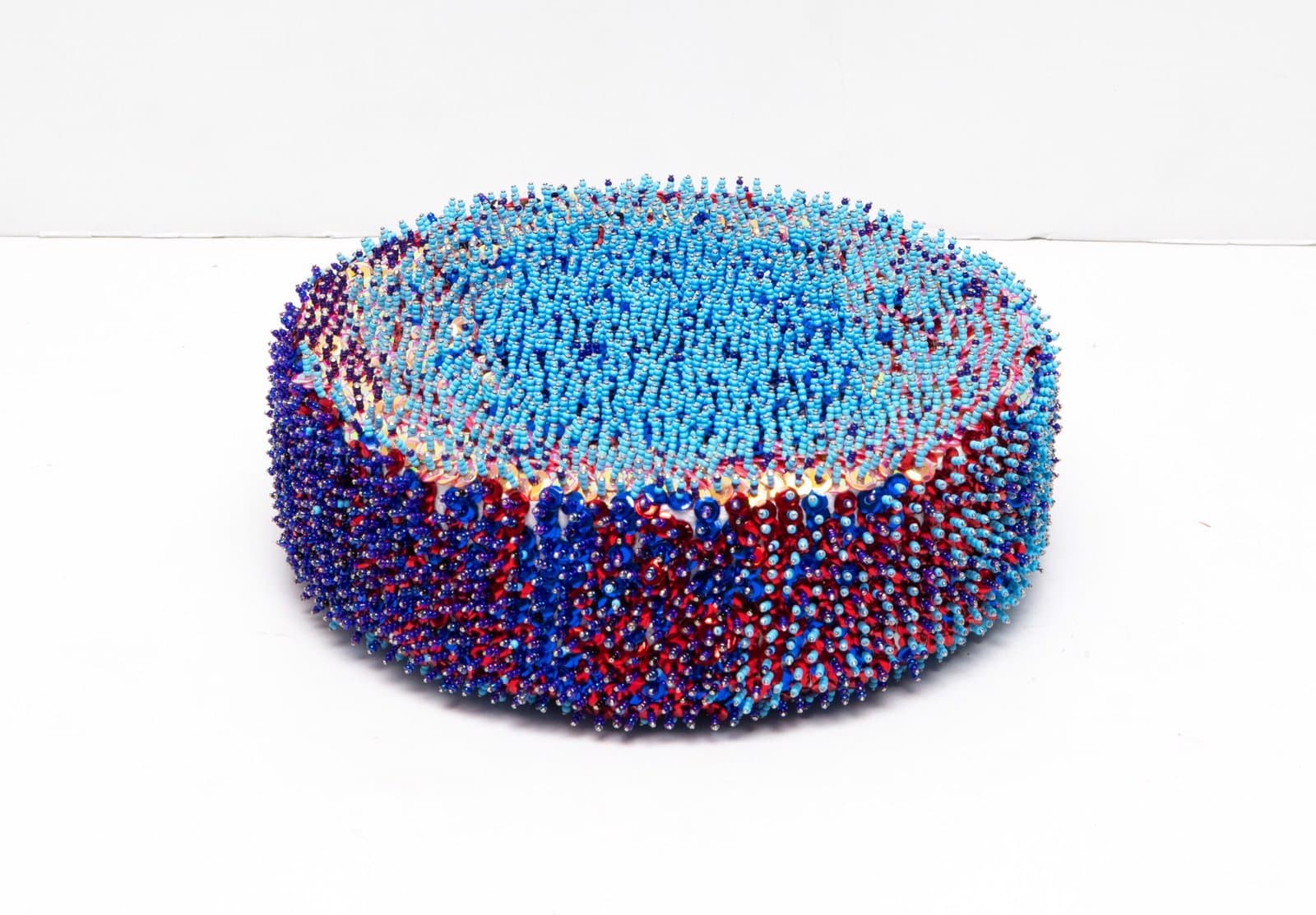 Circular sculptural bowl made of densely packed sequins in blues, reds, and purples on white background.