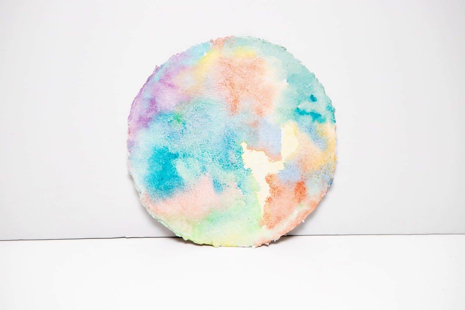Circular watercolor painting with flowing pastel colors including blue, pink, orange, and green on white background
