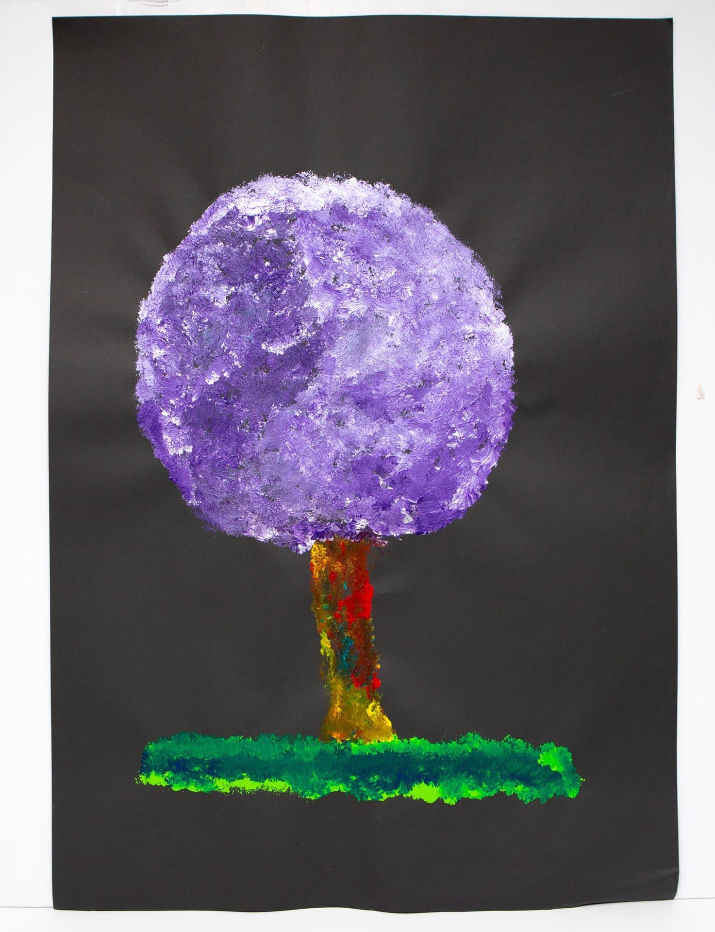 Abstract painting of a tree with purple circular crown, multicolored trunk, and green ground line on dark gray paper.