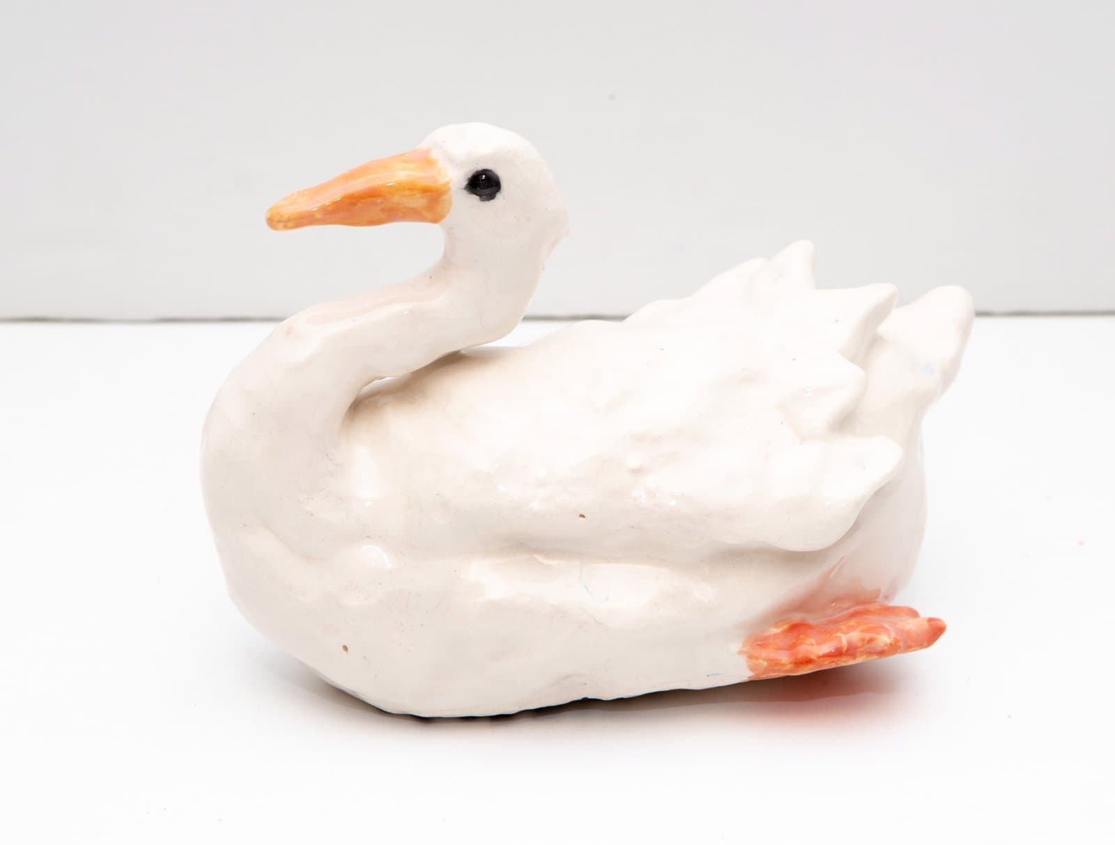 Ceramic swan sculpture in white with orange beak and webbed feet on white surface.