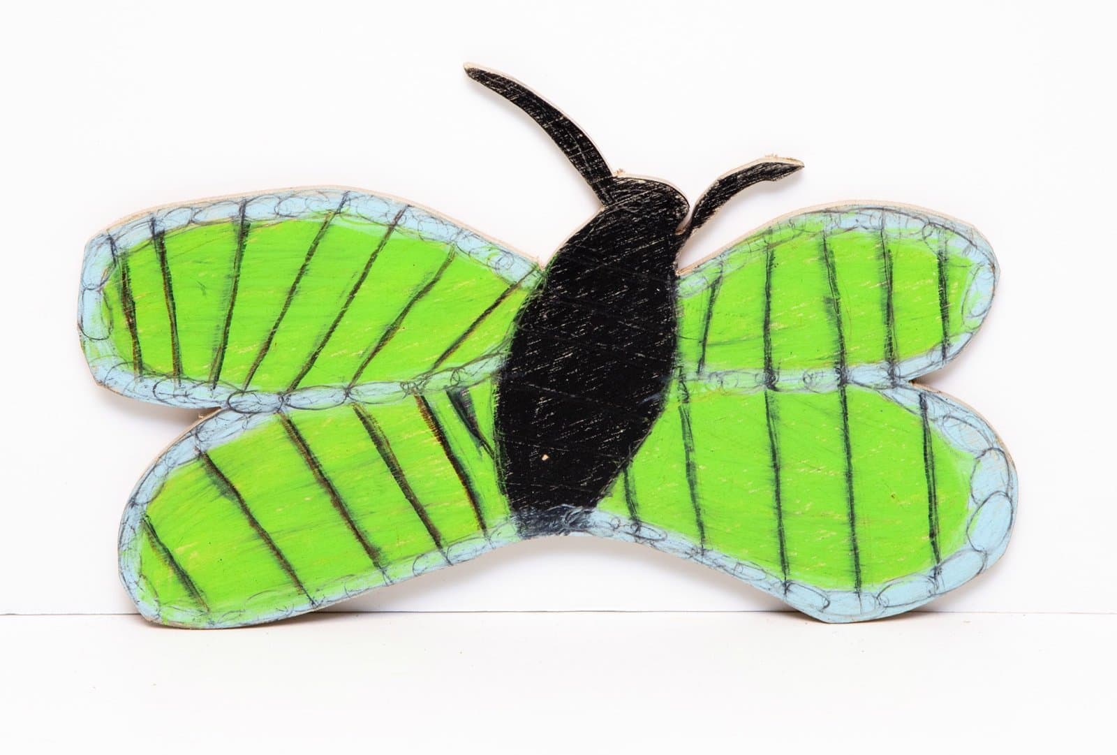 Cut-out butterfly sculpture in colored pencil on wood with bright green wings, black body, and blue borders.