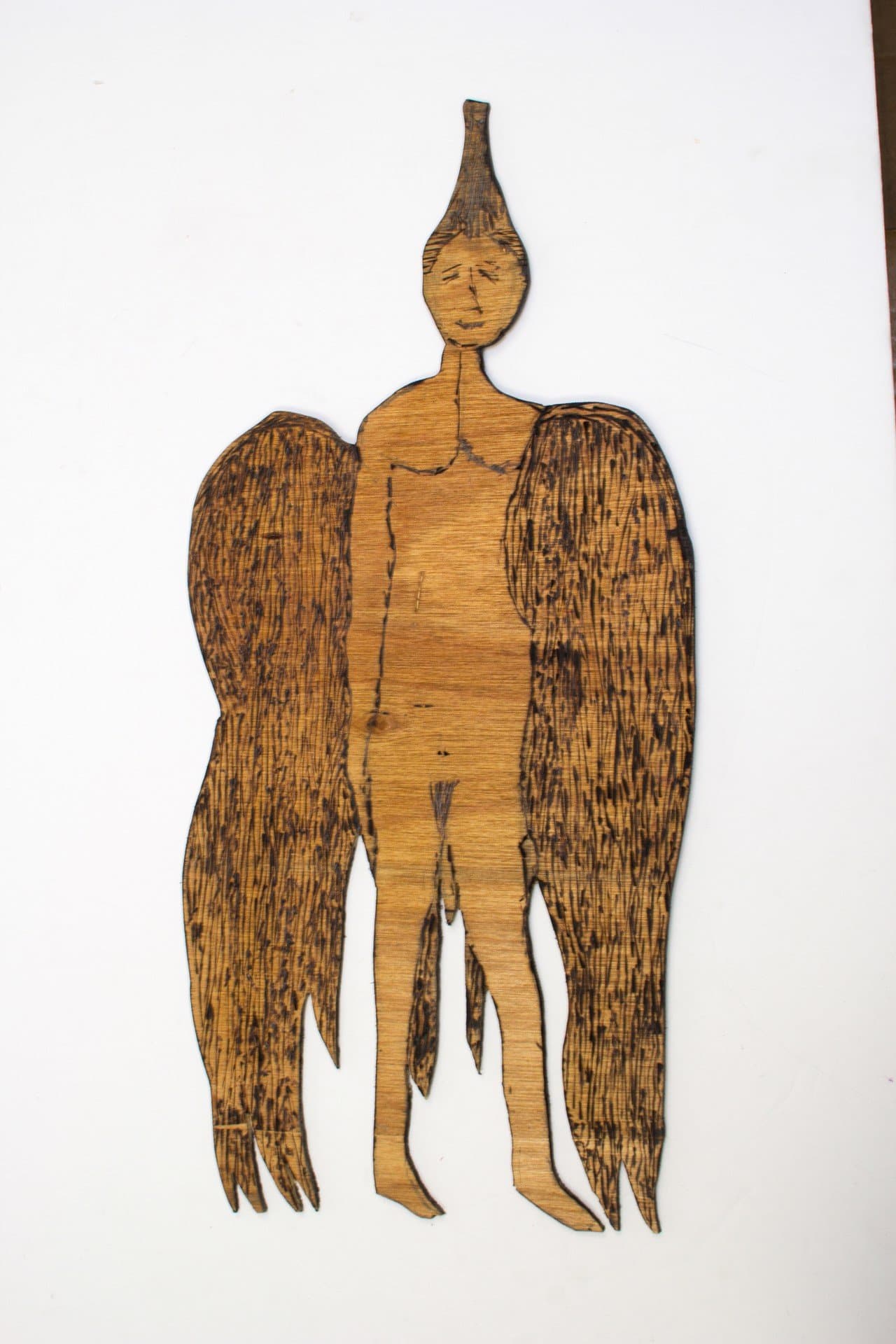 Burned wood sculpture of standing figure with large wings and elongated head against white background