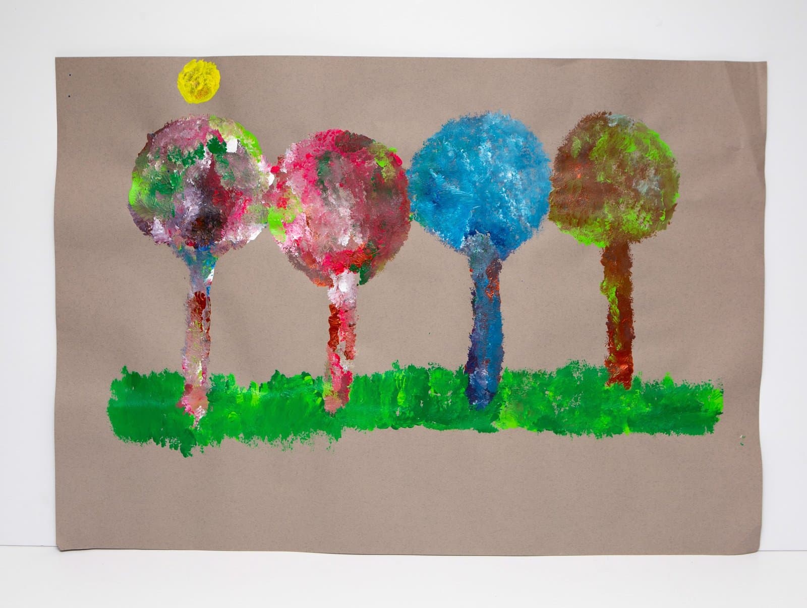 Tempera painting of four colorful trees with circular canopies and a yellow sun on brown paper.