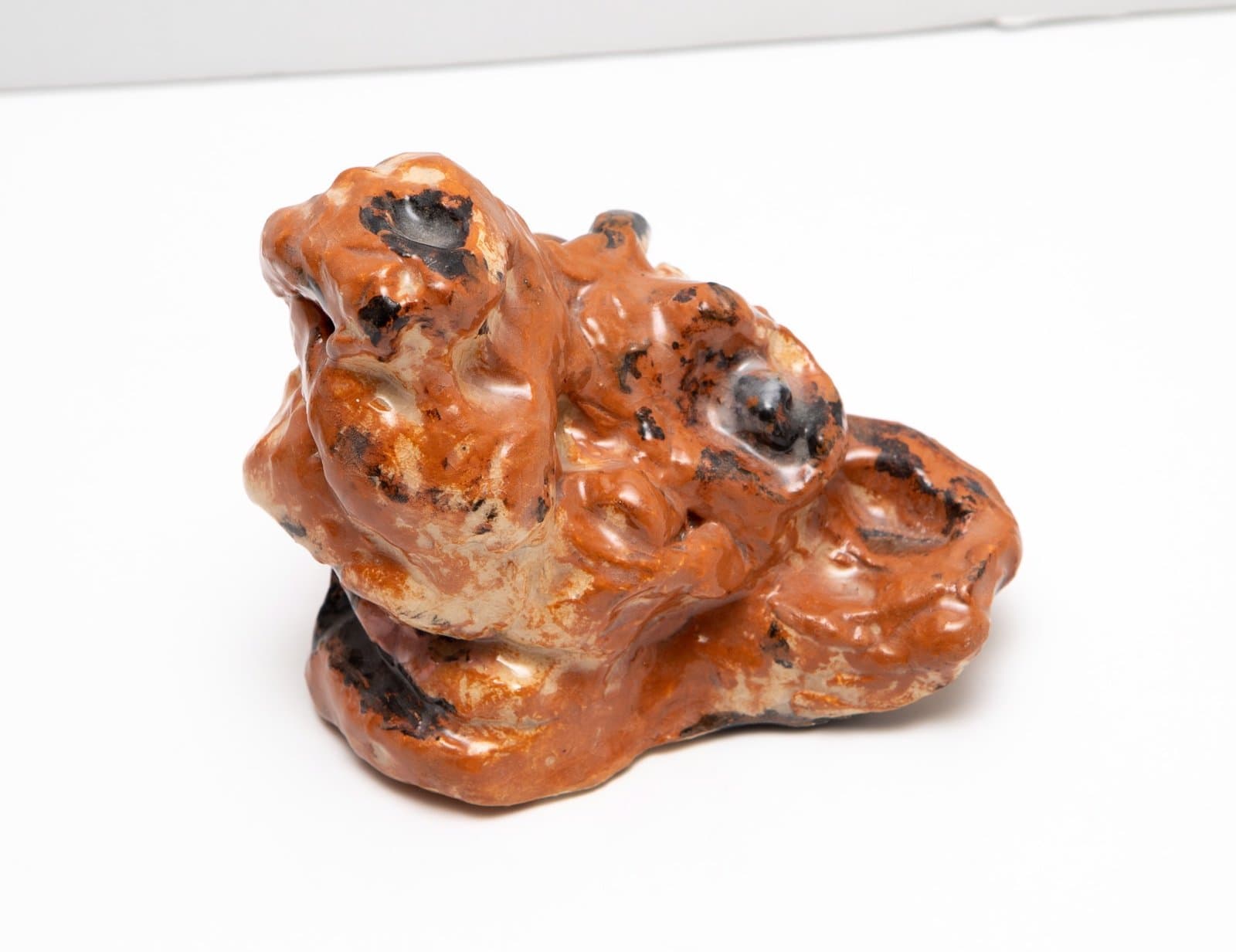 Ceramic sculpture with organic, flowing form in orange-brown glaze with dark spotted accents on white background.