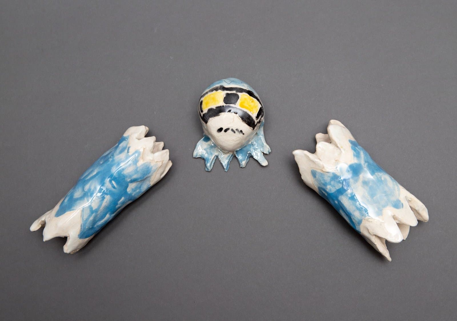 Three ceramic sculptures on gray background: central mask-like head with painted features flanked by two blue and white wing forms