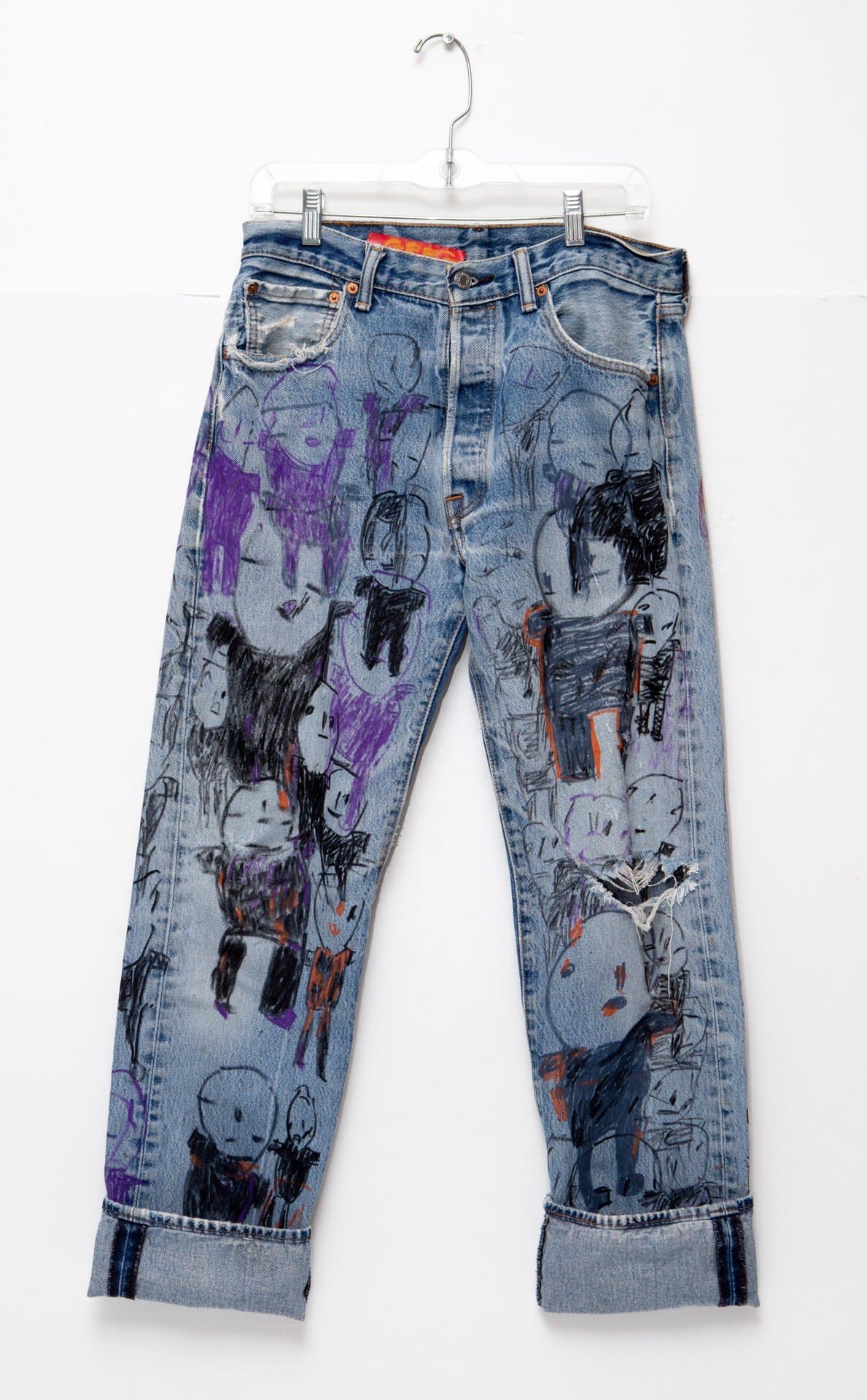 Blue denim jeans decorated with drawings of cartoon-like skull faces in purple, black, and orange marker on hanger