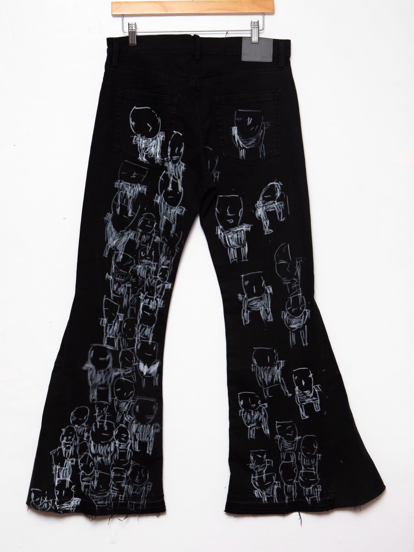 Black denim bell-bottom pants with white drawn figures covering both legs, hanging on wooden hanger against white background.