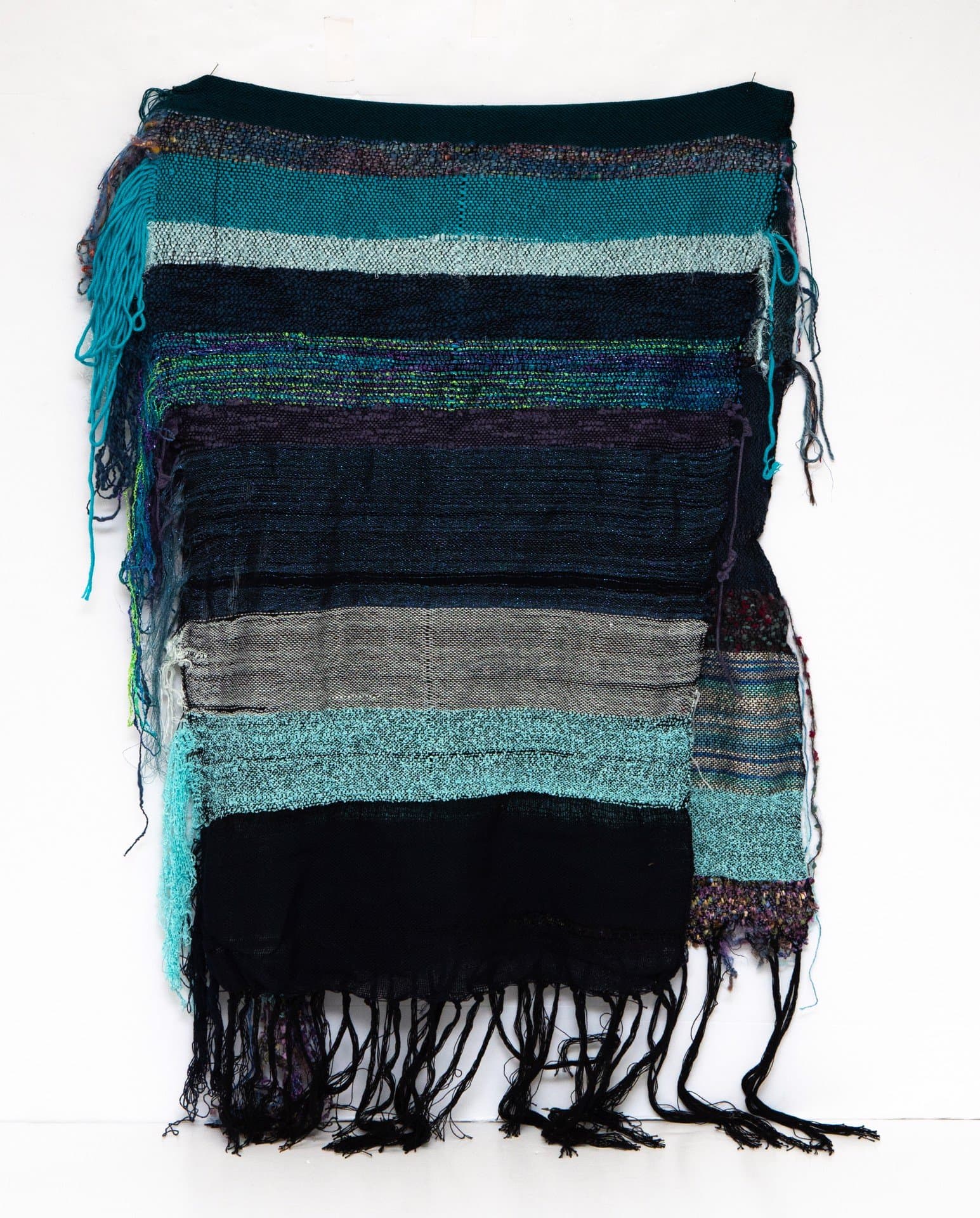 Woven textile with horizontal stripes in turquoise, black, gray, and white with frayed edges and hanging threads.