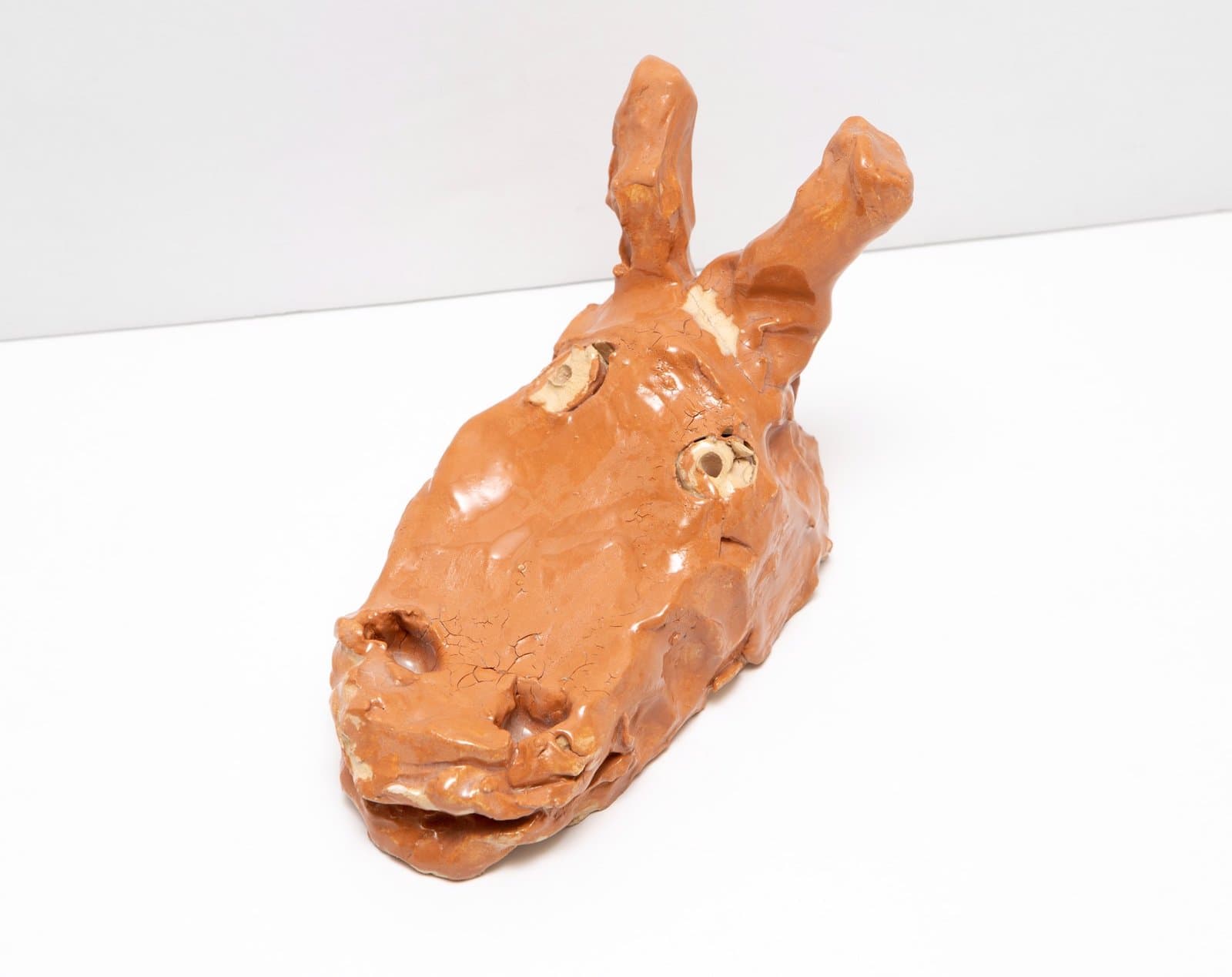 Ceramic sculpture of an orange-brown animal head with upright ears and facial features, displayed on white surface.